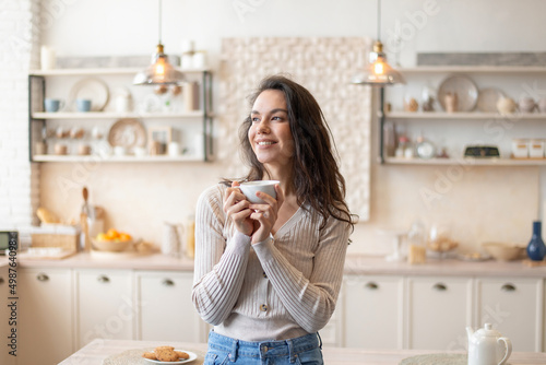Start of the day. Portrait of cheerful woman enjoying morning coffee, holding cup in hands, standing in kitchen