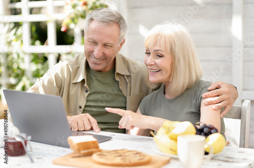 Modern retirement lifestyle concept. Happy senior couple using laptop and browsing internet, sitting outdoors
