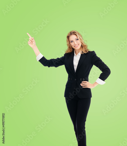 Full body of smiling executive woman in black confident suit, pointing showing copyspace. Business concept. Green background. Young businesswoman.