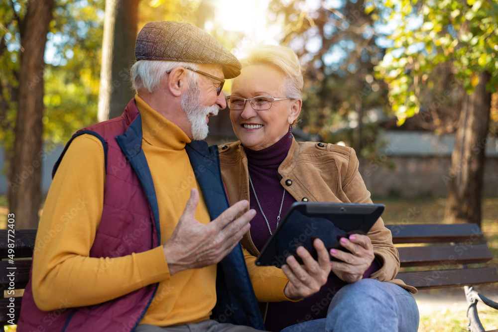 Smiling senior active couple sitting on the bench looking at tablet computer Smiling senior active couple sitting on the bench looking at tablet computer