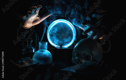 Close-up of the hands of a magician telling the future with a blue crystal ball, surrounded by smoke, books, a skull and mystical elements.