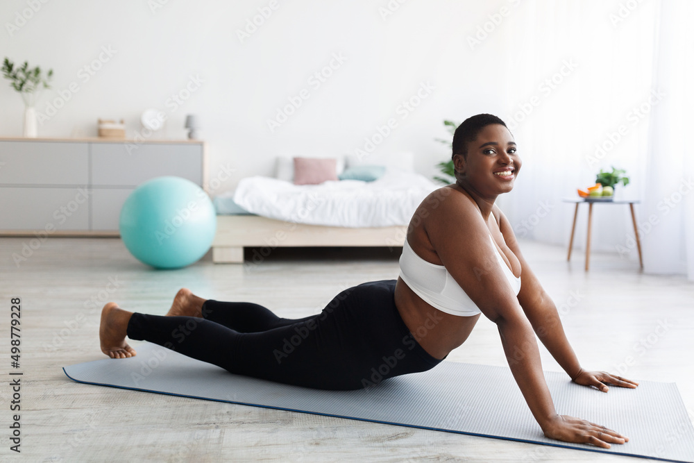 Fototapeta premium Chubby young black lady stretching on sports mat, practicing yoga, trying to lose weight at home, full length