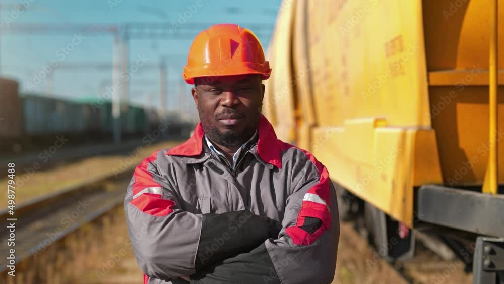 African american railway worker stands at freight train terminal. Railroad man in uniform and red hard hat look at the camera African american railway worker stands at freight train terminal. Railroad man in uniform and red hard hat look at the camera