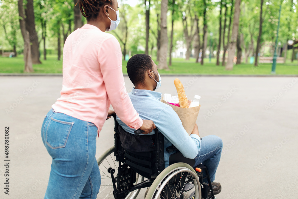 Young black guy in wheelchair holding bag with food, returning home from shopping with his wife during covid lockdown Young black guy in wheelchair holding bag with food, returning home from shopping with his wife during covid lockdown