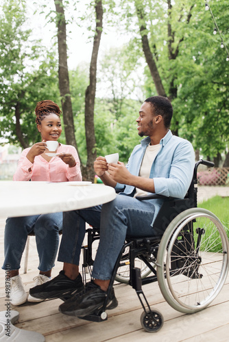 Young African American man in wheelchair having date with lovely woman at outdoor cafe, enjoying coffee at park © Prostock-studio