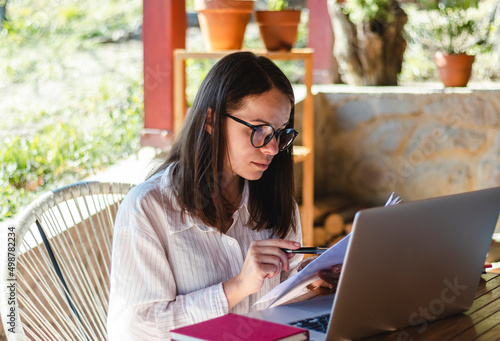 Attractive young woman wearing glasses sits at the table on the terrace and working using her laptop and hard copies of documents.
