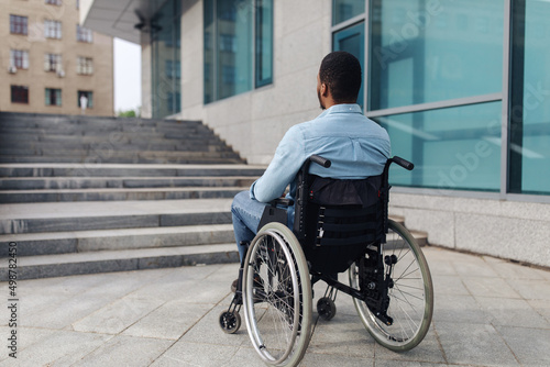 Back view of young black guy in wheelchair standing in front of stairs without ramp, having problem entering building