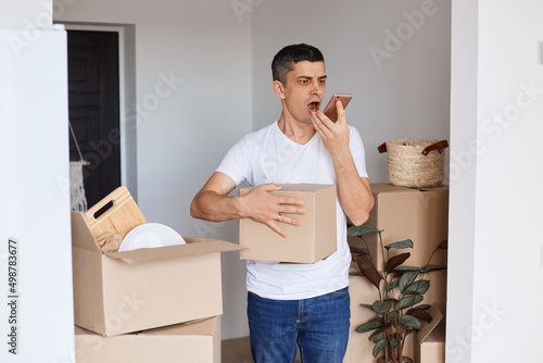 Indoor shot of angry aggressive handsome man wearing white t shirt standing with cardboard box, holding cell phone and screaming with anger while recording voice message.