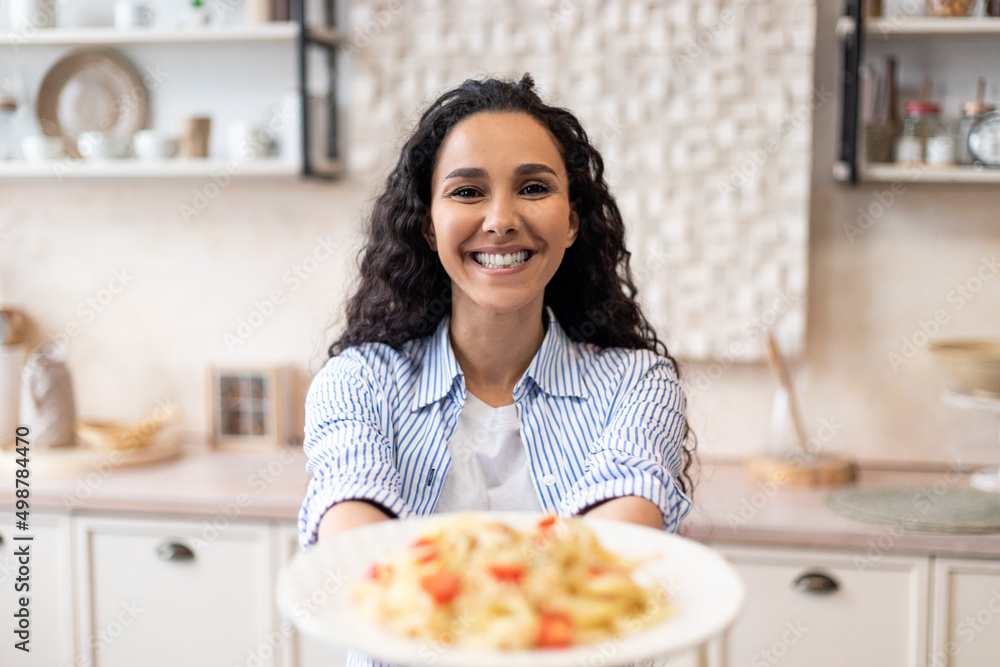 Happy latin woman showing spaghetti in the dish, stretching meal to camera and smiling