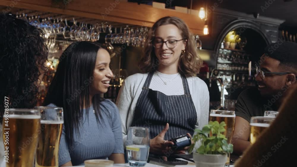 Waitress brings card machine to group of friends at restaurant. One of the women pays for the dinner by tapping mobile phone on card machine. Waitress brings card machine to group of friends at restaurant. One of the women pays for the dinner by tapping mobile phone on card machine.