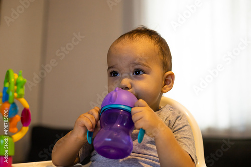 Sweet baby boy sitting in high baby chair and drinking water from baby bottle. Baby at home