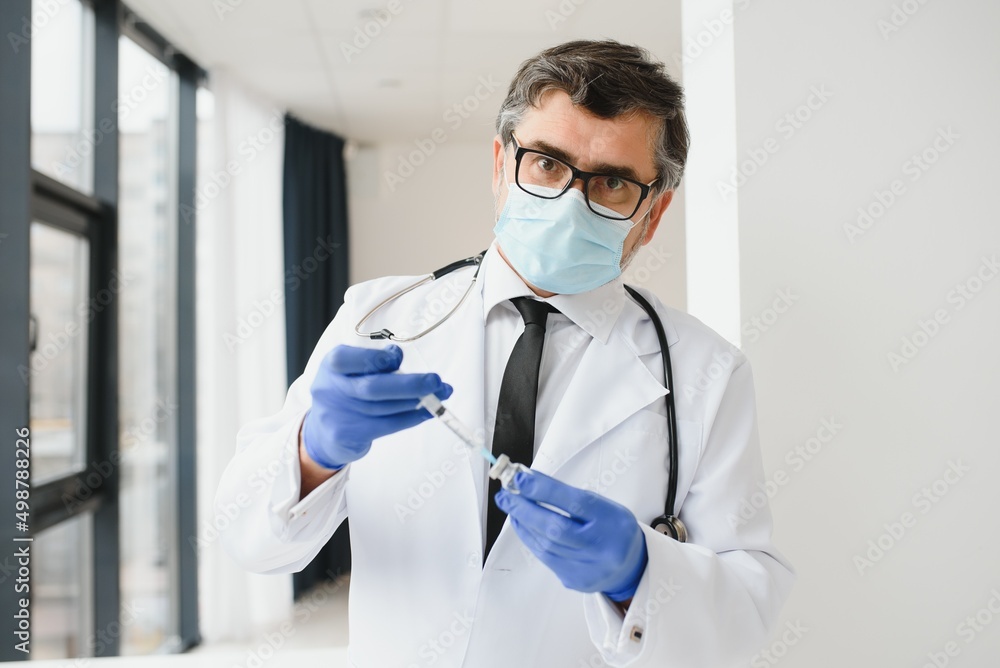Male doctor wearing uniform, mask, medical gloves holding syringe taking covid 19 corona virus vaccine from vial bottle preparing for injection. Coronavirus immunization flu treatment vaccination. Male doctor wearing uniform, mask, medical gloves holding syringe taking covid 19 corona virus vaccine from vial bottle preparing for injection. Coronavirus immunization flu treatment vaccination.