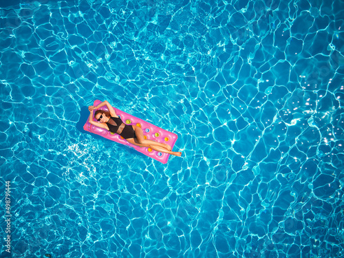 Fit pretty girl in bikini chilling on inflatable pink mattress in swimming pool. Slim hot woman in swimwear tanning. Female relaxing on float in blue water at luxury resort. Aerial, view from above. © artiemedvedev