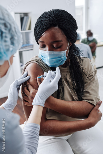 African girl in protective mask getting vaccine in her arm while sitting at hospital during pandemic photo