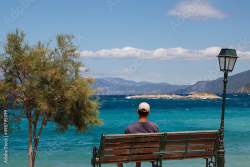 Man looking at the Mediterranean sea and mountains sitting on a bench near a tree and street lamp, Greece