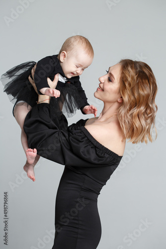 A slender mom lifts the baby up in a black family look, isolated on a gray background.