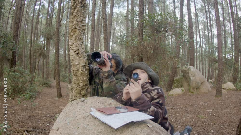 Young mother and son dressed in camouflage clothes photo hunting in forest, staying behind stone. Long-haired woman making wildlife photos while boy observing through spy pipe. Nature, leisure concept Young mother and son dressed in camouflage clothes photo hunting in forest, staying behind stone. Long-haired woman making wildlife photos while boy observing through spy pipe. Nature, leisure concept