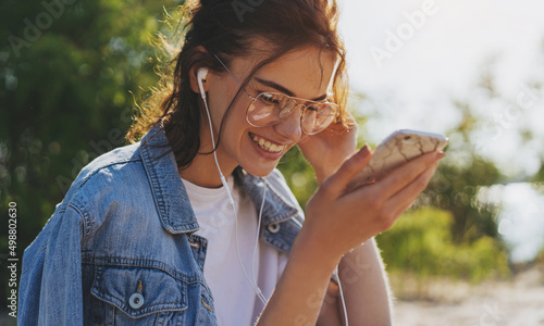 Portrait of happy girl in hipster eyeglasses listening to music while sitting on the beach, cheerful woman speaking with friends outdoors using modern smartphone device