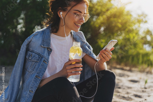 Young positive woman in optical eyeglasses smiling during a video call  on smartphone device, attractive student girl listening to music and reading new on cellphone outdoors