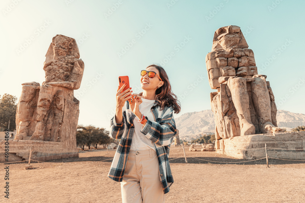 A girl with a smartphone in her hands uses an app or social networks via Wi-Fi Internet standing in front of the famous colossi of Memnon Pharaoh in Luxor