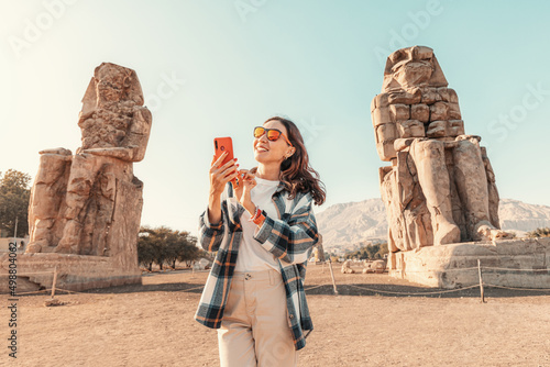 Obraz na płótnie A girl with a smartphone in her hands uses an app or social networks via Wi-Fi Internet standing in front of the famous colossi of Memnon Pharaoh in Luxor w salonie