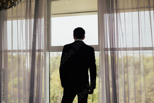 The man in black suit standing near the panoramic window with the green trees background.