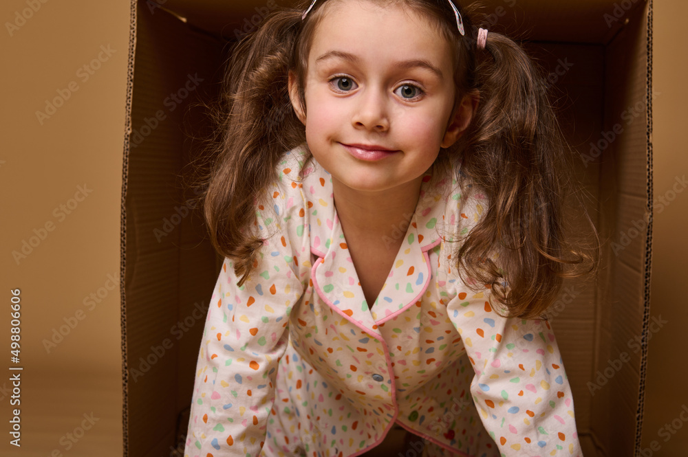 Close-up baby girl with two ponytails in pajamas peeking out from behind a cardboard box, isolated over beige background with copy space Close-up baby girl with two ponytails in pajamas peeking out from behind a cardboard box, isolated over beige background with copy space