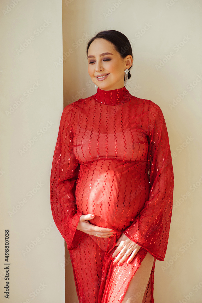 Portrait of attractive pregnant woman in beautiful shiny red dress standing near wall in photostudio and touching her pregnant belly