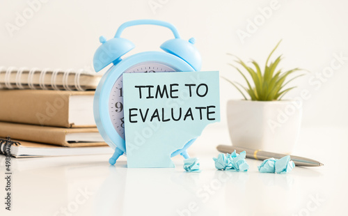 Blue alarm clock, pink, blue and white paper clips, a golden pen and a pink sticker with the text TIME TO EVALUATE