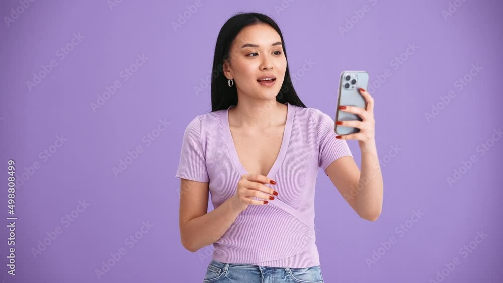 Smiling Asian brunette woman wearing purple t-shirt talking by video call on mobile in the purple studio Smiling Asian brunette woman wearing purple t-shirt talking by video call on mobile in the purple studio