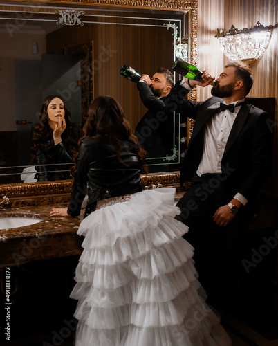 Back view of bride in tiered ruffle wedding dresses and leather black jacket, leaning on washstand and touching her lips while looking to large mirror. Handsome groom in suit, standing and drinking