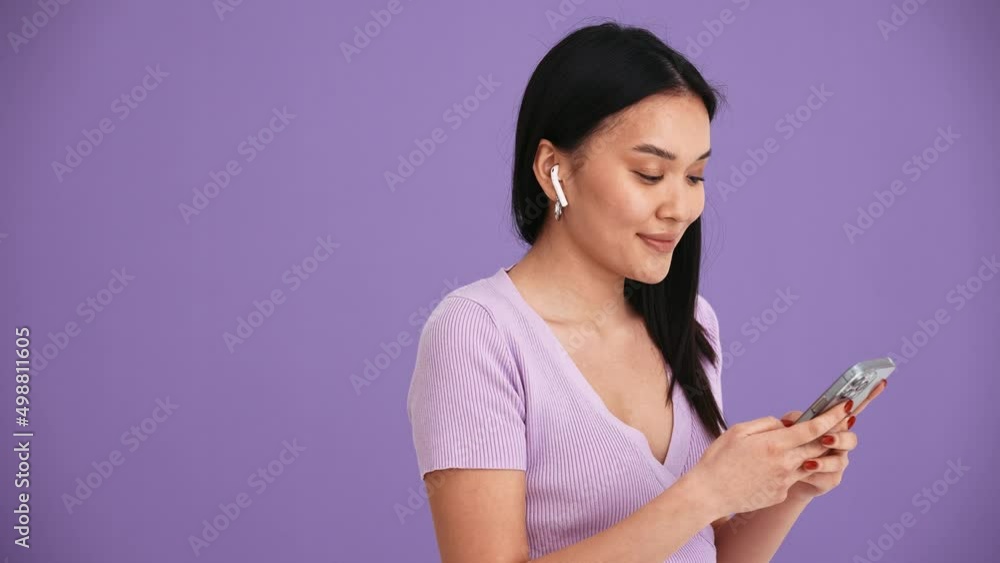 Positive Asian brunette woman in headphones typing by phone in the purple studio
