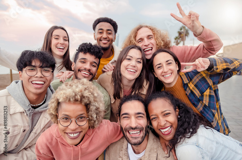 Many beautiful people standing in a circle smile at the camera taking a portrait outdoors - Large group of multiracial friends taking a selfie - Happy young students taking a photo outside school. photo