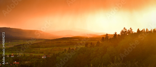 A storm front with heavy rainfall passes over the mountains and the valley, the sun's rays illuminate the landscape.