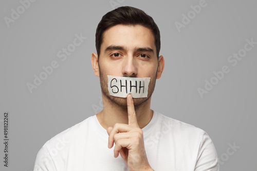 Young man with shhh gesture and taped mouth, asking for silence or to be quiet