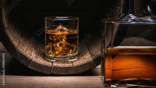 Whiskey with ice in glass stands in wooden barrel of whiskey, next to it is bottle of dark-colored whiskey on close-up wooden table.