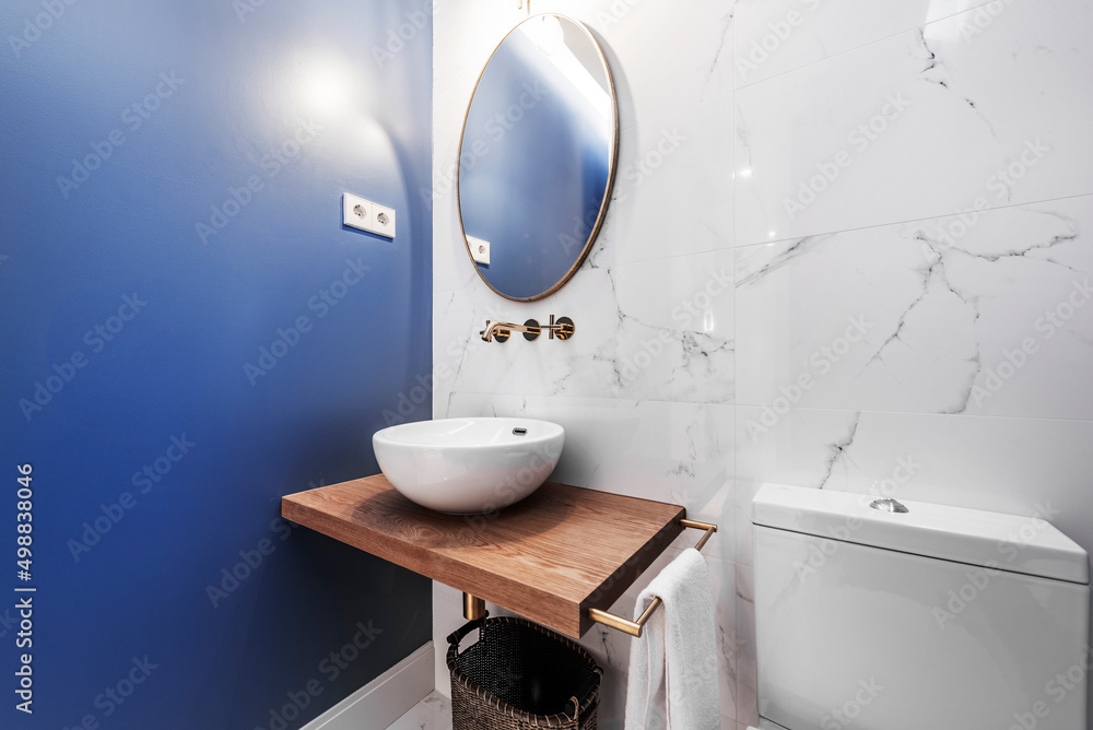 Fototapeta premium Charming bathroom with an oval mirror, a hemispherical porcelain sink on a solid wood countertop, painted blue walls and floors, and another wall covered in white marble with gray veins
