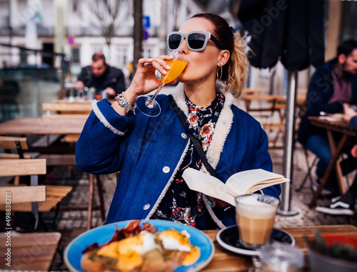 Lifestyle fashion portrait of pretty girl having brunch in outdoors cafe. Drinking an aperitif and enjoying meal. Reading book, enjoying vacation. Wearing stylish jeans coat, sunglasses. Holiday