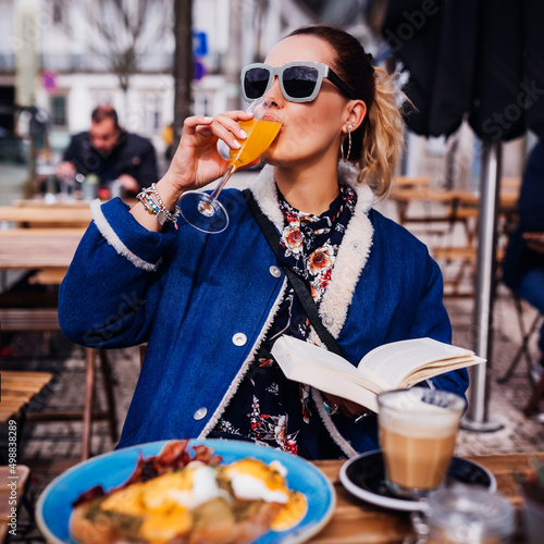 Lifestyle fashion portrait of pretty girl having brunch in outdoors cafe. Drinking an aperitif and enjoying meal. Reading book, enjoying vacation. Wearing stylish jeans coat, sunglasses. Holiday