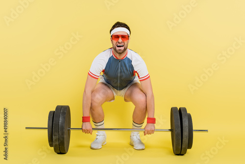 Sportsman screaming while lifting barbell on yellow background.