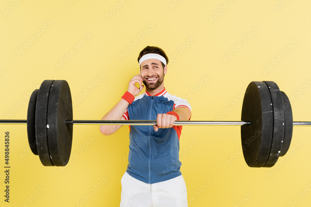 Smiling sportsman talking on smartphone and holding barbell isolated on yellow. Smiling sportsman talking on smartphone and holding barbell isolated on yellow.