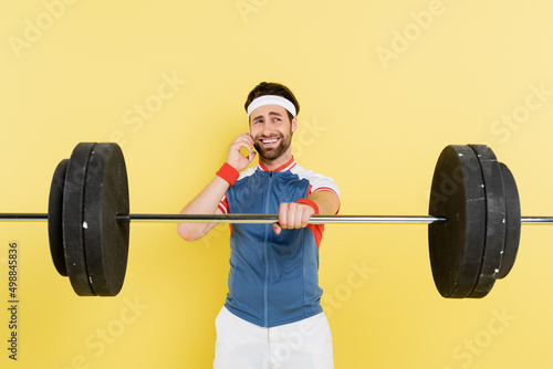 Smiling sportsman talking on smartphone and holding barbell isolated on yellow.