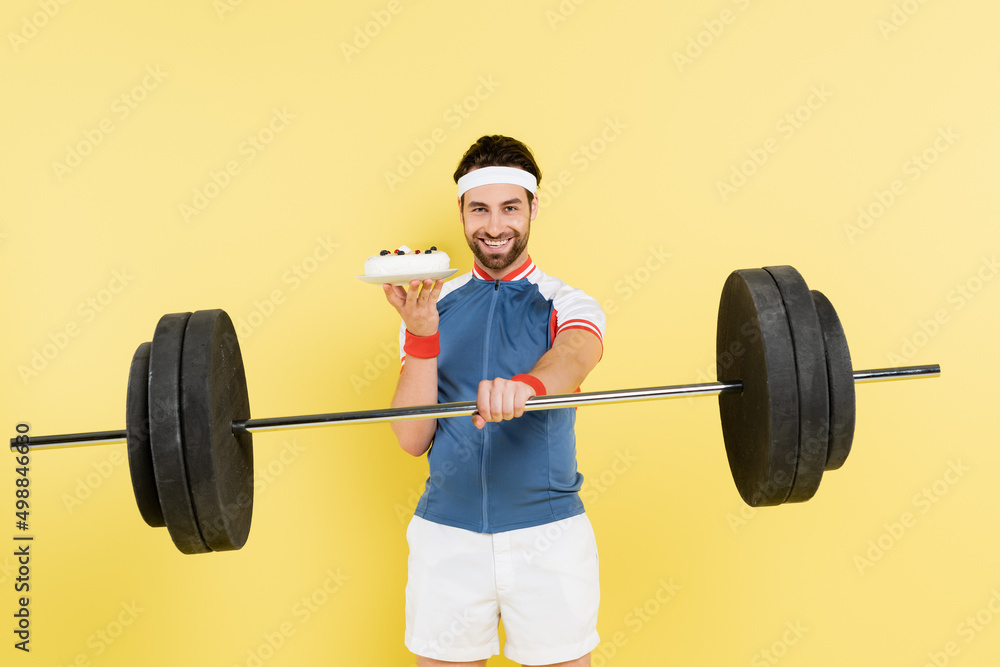 Smiling sportsman holding barbell and cake isolated on yellow. Smiling sportsman holding barbell and cake isolated on yellow.
