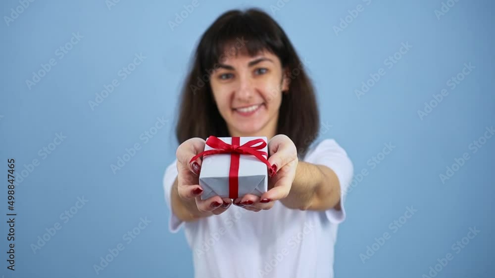 Smiling brunette woman holding gift box and gives it by hands to camera on blue wall background Smiling brunette woman holding gift box and gives it by hands to camera on blue wall background