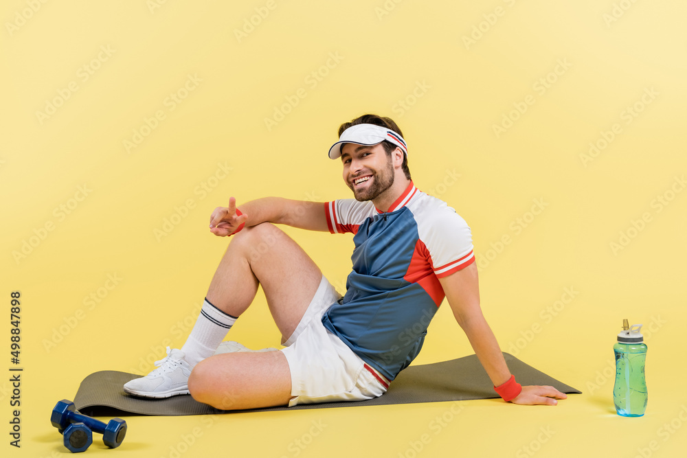 Young sportsman showing peace sign while sitting on fitness mat near dumbbells and sports bottle on yellow background.