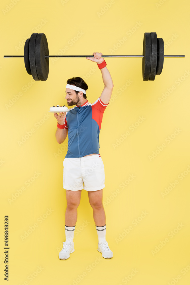 Side view of young sportsman smelling cake and lifting barbell on yellow background. Side view of young sportsman smelling cake and lifting barbell on yellow background.