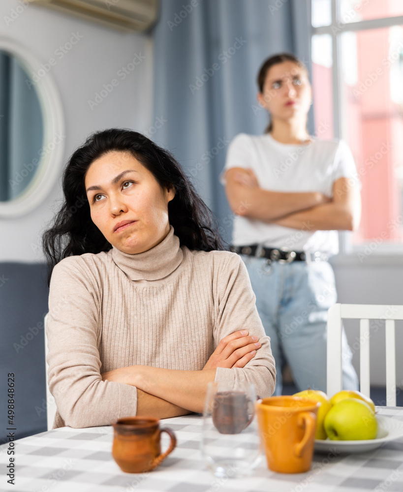 Upset young woman sitting at home table with disgruntled woman behind
