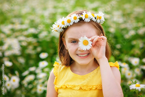 Portrait of a cute village girl in a wreath of daisies in a meadow. He holds a flower in his hands in front of his face. A child on a blooming chamomile field.