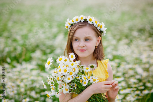 Portrait of a girl in a yellow T-shirt on a blooming field of daisies with a bouquet in her hands and a wreath on her head. A field of daisies on a summer day. Selective focus.