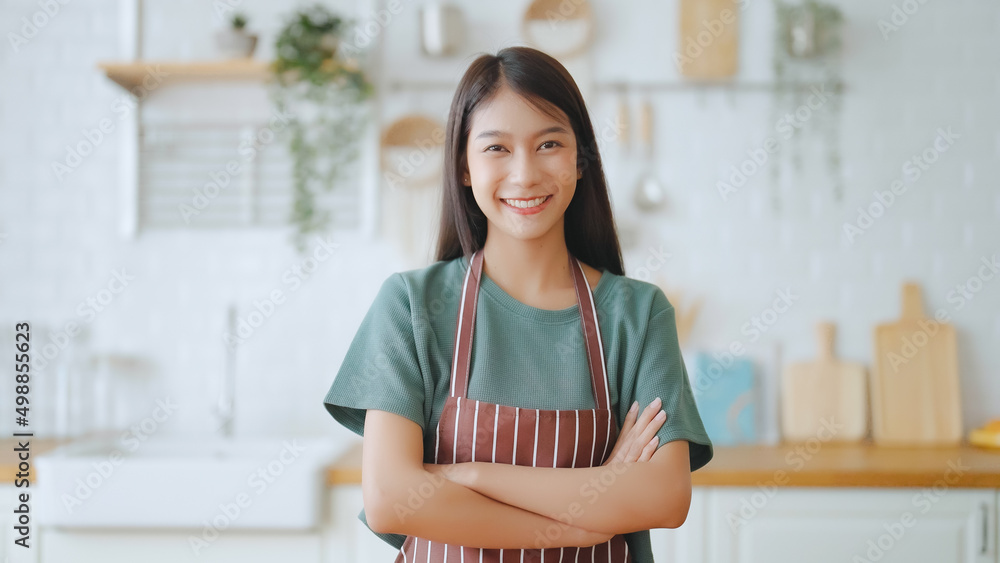 Happy young asian woman wearing apron and standing in kitchen room. Beautiful female smiling and looking at camera feeling confident Happy young asian woman wearing apron and standing in kitchen room. Beautiful female smiling and looking at camera feeling confident